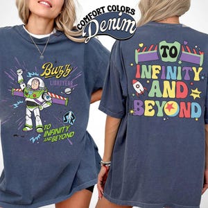 Op de afbeelding: Denimblauwe t-shirt met Buzz Lightyear uit Toy Story. De voorkant heeft Buzz in zijn ruimtepak met de woorden "Buzz Lightyear" en "To Infinity and Beyond". De achterkant van de shirt heeft de zin "To Infinity and Beyond" in kleurrijke letters.