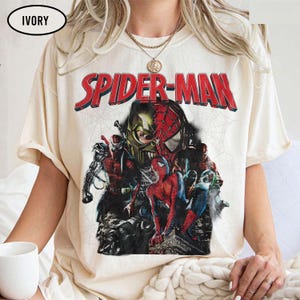 Pode incluir: Uma t-shirt de manga curta na cor marfim com uma grande estampa gr&aacute;fica de personagens do Spider-Man. O texto SPIDER-MAN aparece em letras vermelhas em negrito sobre uma colagem do her&oacute;i em v&aacute;rias poses e um design de m&aacute;scara sobre teia.