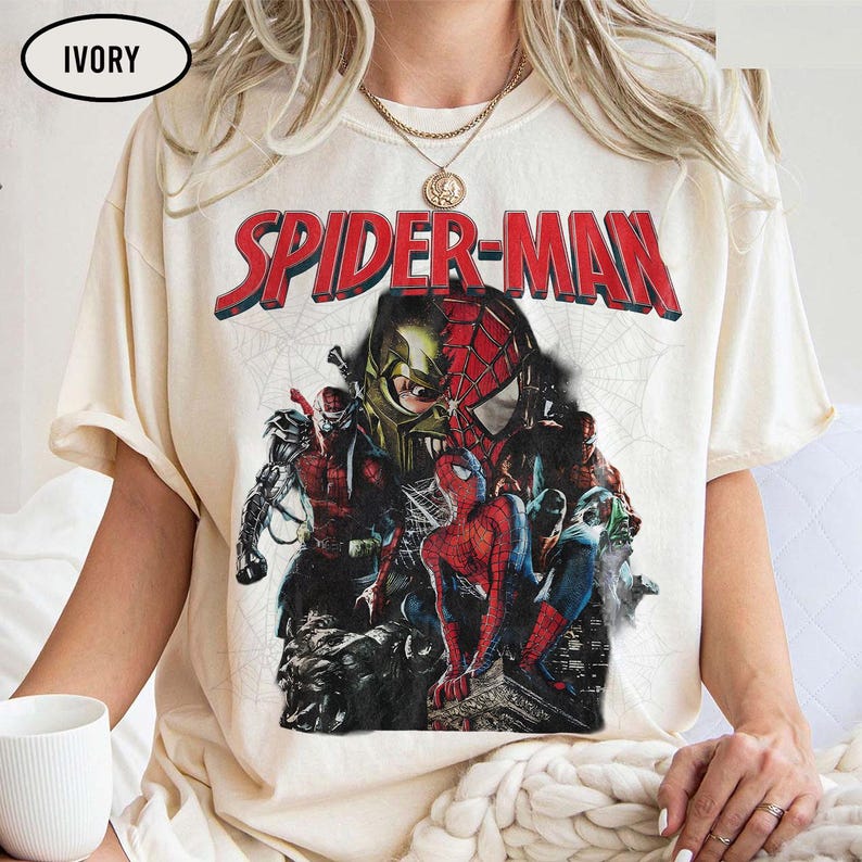 Pode incluir: Uma t-shirt de manga curta na cor marfim com uma grande estampa gr&aacute;fica de personagens do Spider-Man. O texto SPIDER-MAN aparece em letras vermelhas em negrito sobre uma colagem do her&oacute;i em v&aacute;rias poses e um design de m&aacute;scara sobre teia.
