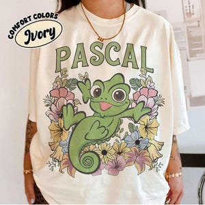 May include: An ivory t-shirt featuring a graphic of a green chameleon surrounded by colorful flowers. The text PASCAL is printed in green above the chameleon. A circular logo in the top left corner contains the text COMFORT COLORS and IVORY.