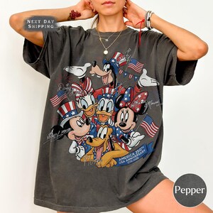 May include: Dark grey t-shirt with a graphic of Disney characters Mickey Mouse, Minnie Mouse, Donald Duck, Daisy Duck, Goofy, and Pluto, celebrating the USA's 250th anniversary. The design includes American flags and patriotic colours.