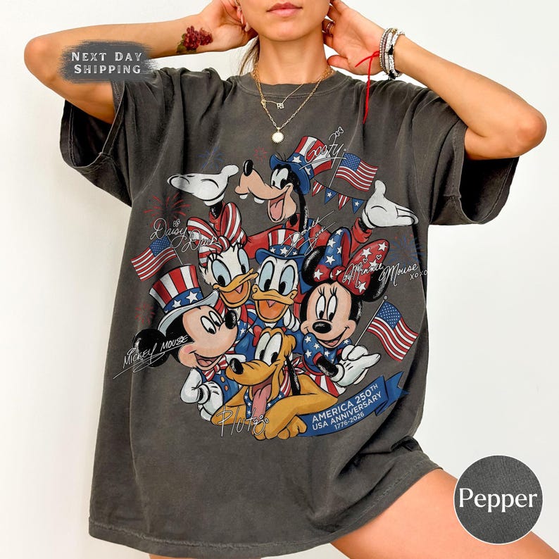 May include: Dark grey t-shirt with a graphic of Disney characters Mickey Mouse, Minnie Mouse, Donald Duck, Daisy Duck, Goofy, and Pluto, celebrating the USA's 250th anniversary. The design includes American flags and patriotic colours.