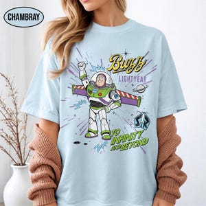May include: Light blue t-shirt featuring a graphic of Buzz Lightyear with the text "Buzz Lightyear" and "To Infinity and Beyond." The design includes a space theme with a planet and a rocket ship. The shirt has a crew neck and short sleeves.