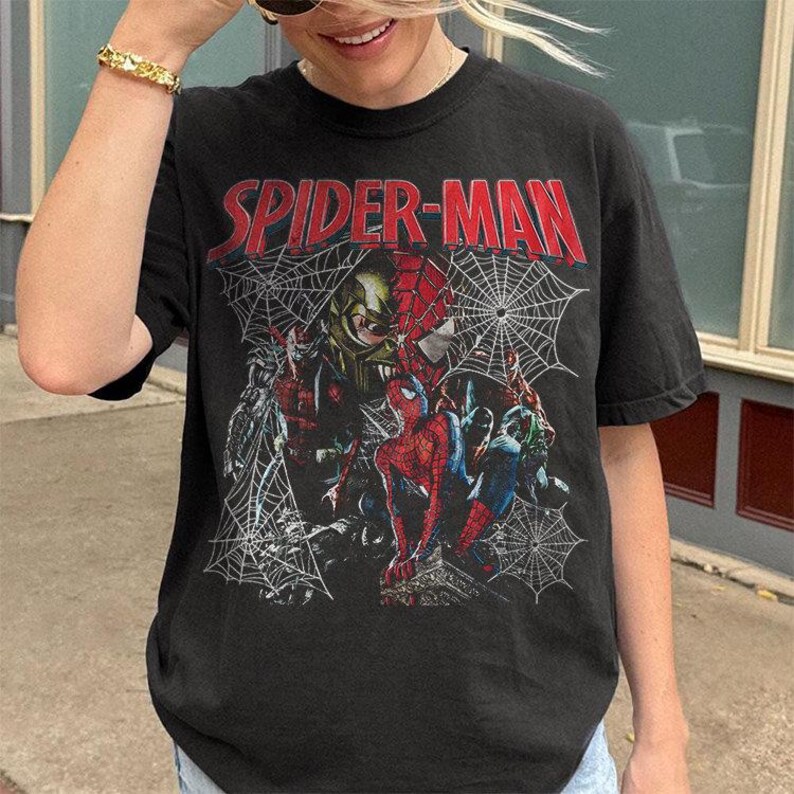 Pode incluir: Uma t-shirt preta de manga curta com uma estampa gr&aacute;fica do Spider-Man e do Duende Verde. O texto SPIDER-MAN est&aacute; impresso a vermelho no topo. O design inclui padr&otilde;es de teia de aranha brancos e v&aacute;rias figuras num estilo de colagem.