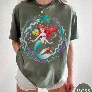 May include: Moss green t-shirt with a vibrant graphic of a mermaid with red hair, a blue tail, and a purple seashell bra. The design includes a yellow fish, a red crab, and aquatic plants. The word "MOSS" is in a circle at the bottom right.