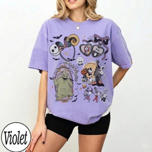 May include: Violet-colored t-shirt featuring a collage of cartoon characters and Halloween-themed graphics. The design includes images of a skeleton, a heart, a ghost, and other spooky elements. The word "Nightmare" is also visible.