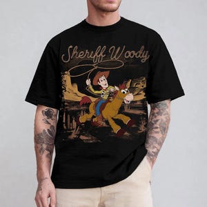 K&ouml;nnte beinhalten: Schwarzes T-Shirt mit einer Cartoon-Illustration von Sheriff Woody auf einem Pferd mit Lasso. Der Text "Sheriff Woody" steht in einer strukturierten braunen Schrift &uuml;ber dem Bild. Das Design ist vor einer W&uuml;stenlandschaft.