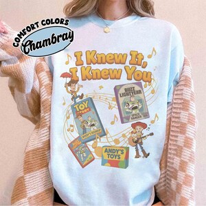 K&ouml;nnte beinhalten: Ein hellblaues Sweatshirt mit Rundhalsausschnitt und einem Grafikdruck mit dem gelben Text "I Knew It, I Knew You". Das Design zeigt Illustrationen von Woody, Buzz Lightyear und Jessie aus Toy Story sowie Noten und Spielzeugschachteln.