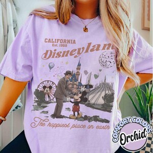 Puede incluir: Una camiseta de color lila con un gr&aacute;fico de Disneyland. El gr&aacute;fico presenta a Mickey Mouse, un hombre con traje, una noria y un castillo. El texto incluye "CALIFORNIA Est. 1955", "Disneyland" y "The happiest place on earth".