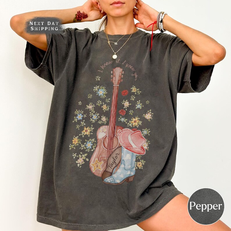 May include: A charcoal gray short-sleeve t-shirt featuring a graphic print of an acoustic guitar, a cowboy boot, and a cowboy hat surrounded by small colorful flowers. The text "I knew you" is printed in a curved arc above the guitar illustration.