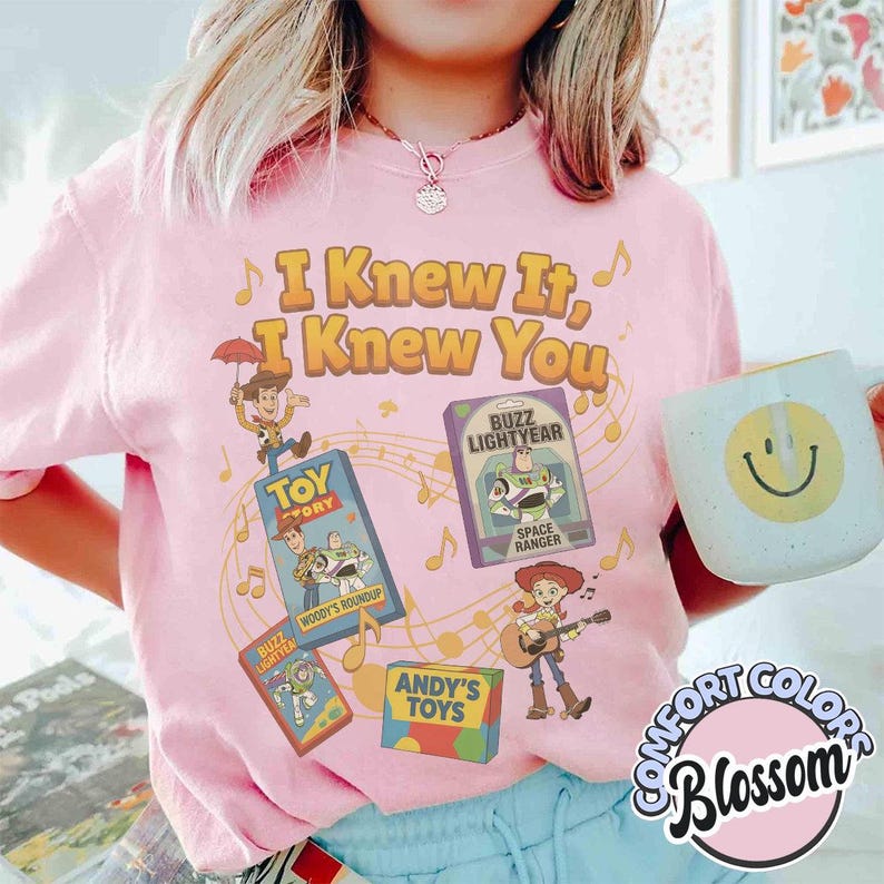 K&ouml;nnte beinhalten: Ein hellrosa T-Shirt mit dem Text I Knew It, I Knew You und bunten Grafiken von Toy Story-Figuren, darunter Woody und Buzz Lightyear, sowie Noten. Ein heller Becher mit einem gelben Smiley-Gesicht ist ebenfalls zu sehen.