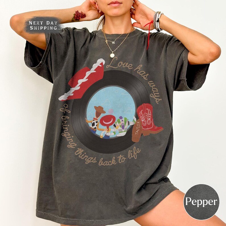 May include: A charcoal gray short-sleeve t-shirt featuring a graphic of a black vinyl record with a central illustration of three cartoon characters. The text reads, Love has ways of bringing things back to life, with a red cowboy boot and hat.