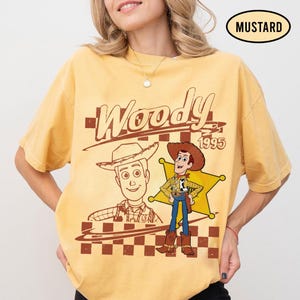 May include: Mustard-colored t-shirt featuring a graphic of Woody from Toy Story. The design includes the word "Woody" in a vintage font, the year "1995", and an illustration of Woody in his cowboy attire. A checkered pattern is at the bottom.