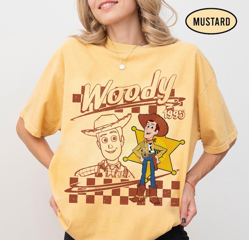 May include: Mustard-colored t-shirt featuring a graphic of Woody from Toy Story. The design includes the word "Woody" in a vintage font, the year "1995", and an illustration of Woody in his cowboy attire. A checkered pattern is at the bottom.