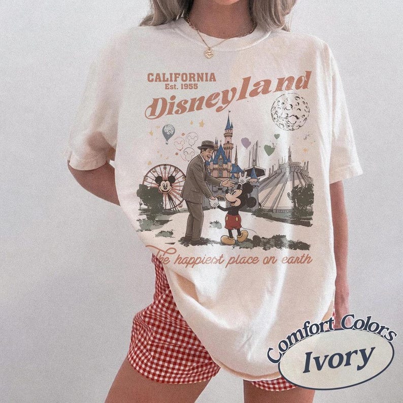 May include: Ivory-colored t-shirt with a Disneyland graphic. The graphic features Mickey Mouse, a man in a suit, and iconic park imagery. Text includes "California Disneyland Est. 1955" and "The happiest place on earth."