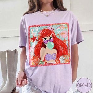 May include: A light purple t-shirt featuring a colorful graphic of a mermaid with red hair, heart-shaped sunglasses, and a bubble. The graphic is framed with a red border and surrounded by sea creatures.