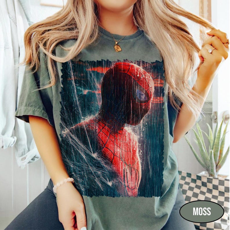 Pu&ograve; includere: T-shirt verde muschio con grafica di Spider-Man in una scena piovosa. Il costume rosso e nero del supereroe si staglia su uno sfondo scuro e strutturato. La maglietta ha un girocollo classico e maniche corte.