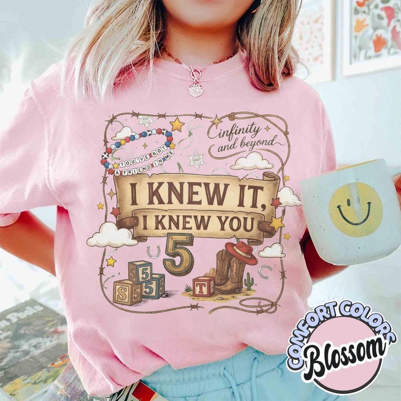 May include: A light pink t-shirt featuring a graphic print with a brown scroll banner that reads I KNEW IT, I KNEW YOU, the number 5, a cowboy boot, alphabet blocks, and the text Cinfinity and beyond. A light blue mug with a yellow smiley face is held.