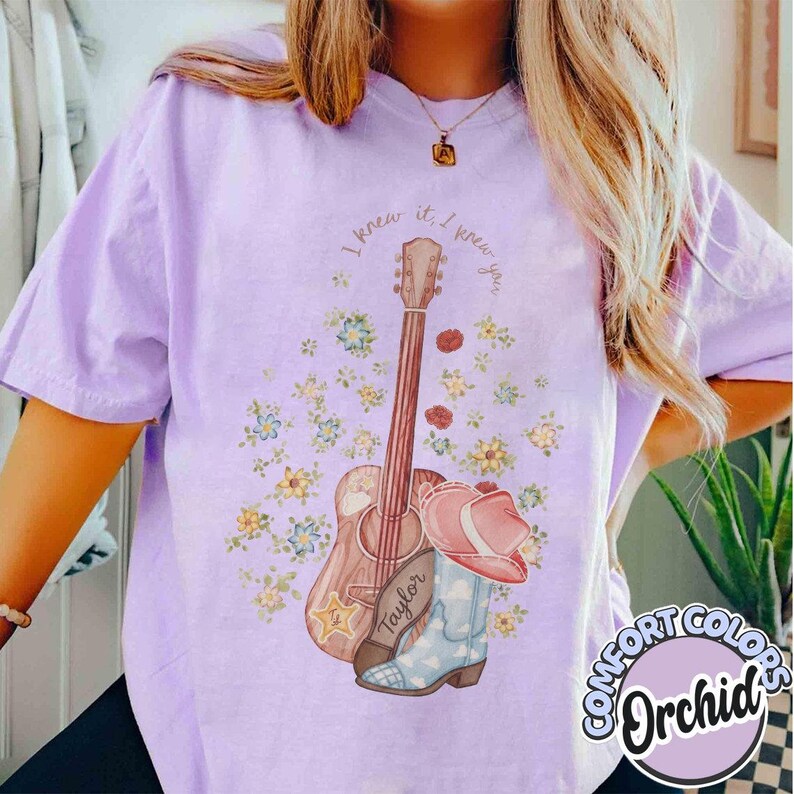 May include: A light purple t-shirt featuring a graphic print of an acoustic guitar, a pink cowboy hat, and a light blue cowboy boot surrounded by small yellow and blue flowers. The text "I knew it, I knew you" is printed in a curved arc above.
