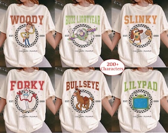 May include: A collection of six cream-colored t-shirts featuring graphic prints of cartoon characters including Woody, Buzz Lightyear, Slinky, Forky, Bullseye, and Lilypad. Each shirt displays the character name, the text EST. 1999, and custom name.
