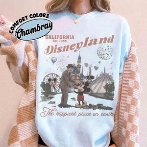 Puede incluir: Sudadera azul claro de Disneyland con un gr&aacute;fico de estilo vintage. El dise&ntilde;o presenta a Mickey Mouse, un hombre con traje, una noria y el texto "Disneyland California Est. 1955" y "The happiest place on earth."