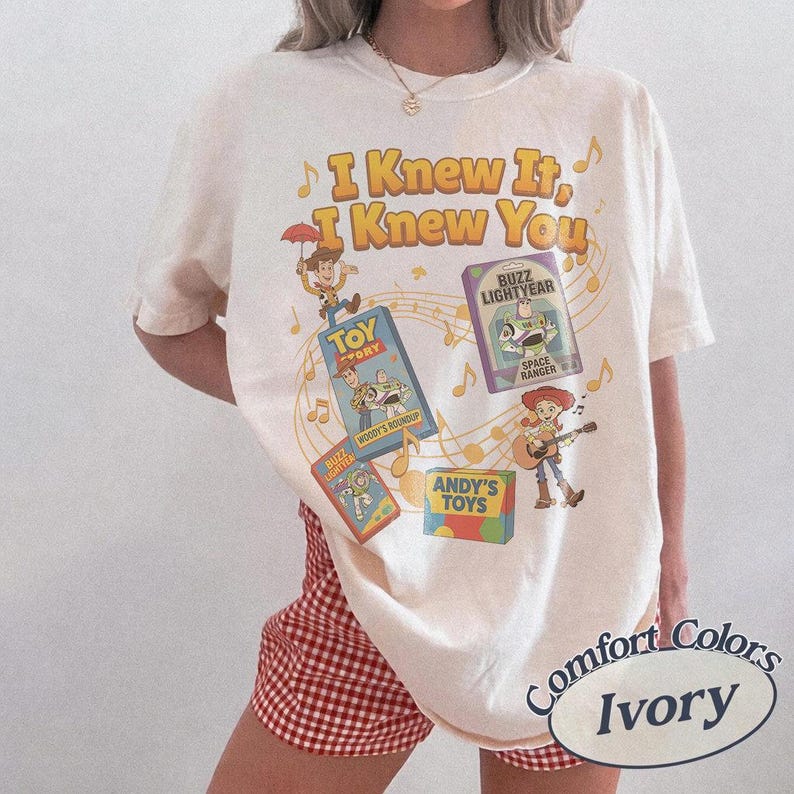K&ouml;nnte beinhalten: Ein elfenbeinfarbenes Kurzarm-T-Shirt mit einem Grafikdruck und dem Text I Knew It, I Knew You. Das Design zeigt Illustrationen von Woody, Buzz Lightyear und Jessie aus Toy Story sowie Noten und verschiedene Spielzeugverpackungen.