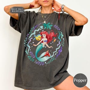 May include: Dark grey t-shirt with a vibrant illustration of a mermaid with red hair, a blue tail, and a purple seashell bra. The design includes a yellow fish, a red crab, and other sea elements. The shirt has the text "Next Day Shipping" on the top left and "Pepper" on the bottom right.