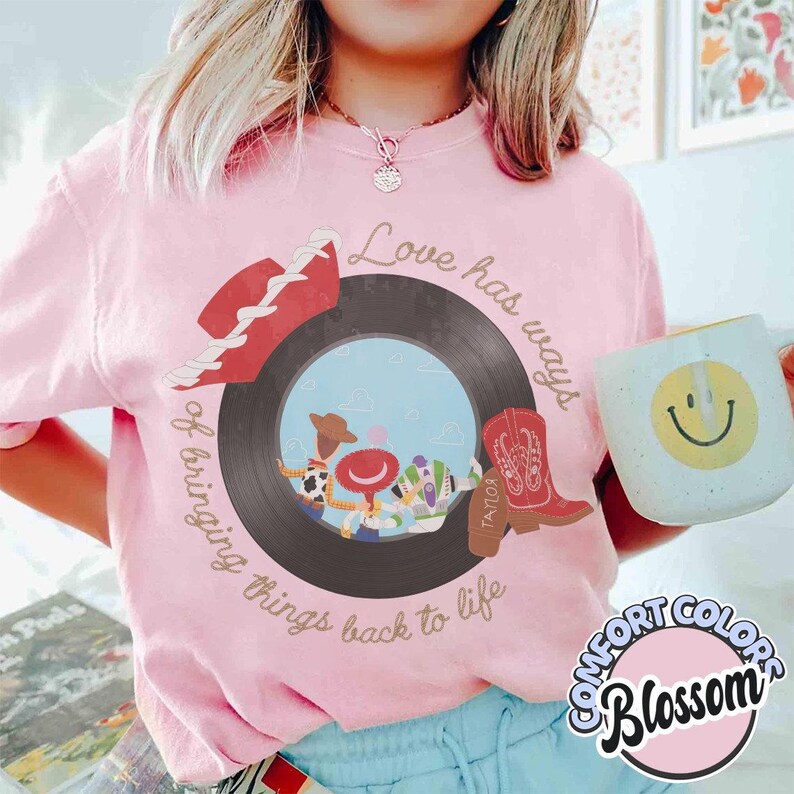 May include: A light pink t-shirt featuring a graphic of a black vinyl record with a blue sky center. The design includes a red cowboy hat, a red cowboy boot, and three cartoon characters. The text reads, Love has ways of bringing things back to life.