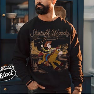 K&ouml;nnte beinhalten: Schwarzes Sweatshirt mit einer Grafik von Sheriff Woody auf einem Pferd und dem Text "Sheriff Woody". Das Sweatshirt hat einen Rundhalsausschnitt und lange &Auml;rmel. Die Worte "Comfort Colors Black" sind links unten aufgedruckt.