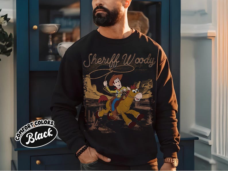 K&ouml;nnte beinhalten: Schwarzes Sweatshirt mit einer Grafik von Sheriff Woody auf einem Pferd und dem Text "Sheriff Woody". Das Sweatshirt hat einen Rundhalsausschnitt und lange &Auml;rmel. Die Worte "Comfort Colors Black" sind links unten aufgedruckt.