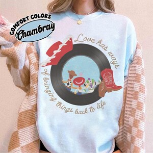 May include: A light blue crewneck sweatshirt featuring a graphic of a black vinyl record with cartoon characters inside the center hole. The design includes a red cowboy hat, a red cowboy boot, and the text Love has ways of bringing things back to life.