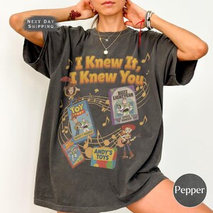 K&ouml;nnte beinhalten: Ein anthrazitfarbenes Kurzarm-T-Shirt mit dem Schriftzug I Knew It, I Knew You in goldener Schrift. Die Grafik enth&auml;lt Illustrationen der Toy Story-Figuren Buzz Lightyear, Woody und Jessie sowie Noten und Spielzeugschachteln.