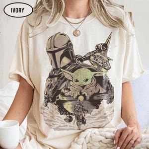 May include: Ivory-colored t-shirt featuring a graphic of The Mandalorian and Grogu. The design includes a spaceship and the Mandalorian's helmet. The shirt is a light cream color and has short sleeves.