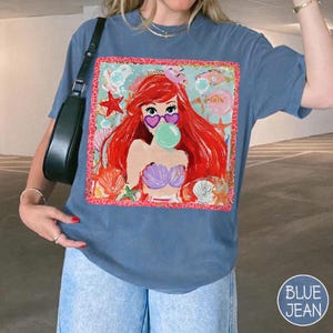May include: A blue t-shirt featuring a graphic of a mermaid with red hair, heart-shaped sunglasses, and a bubble. The graphic is framed with a pink border and surrounded by seashells and starfish. The shirt has short sleeves.