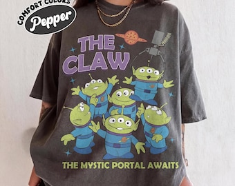 May include: A charcoal gray short-sleeve t-shirt featuring a graphic of five green three-eyed aliens in blue space suits. The text reads THE CLAW and THE MYSTIC PORTAL AWAITS. A small planet graphic and a mechanical claw are also printed on the front.