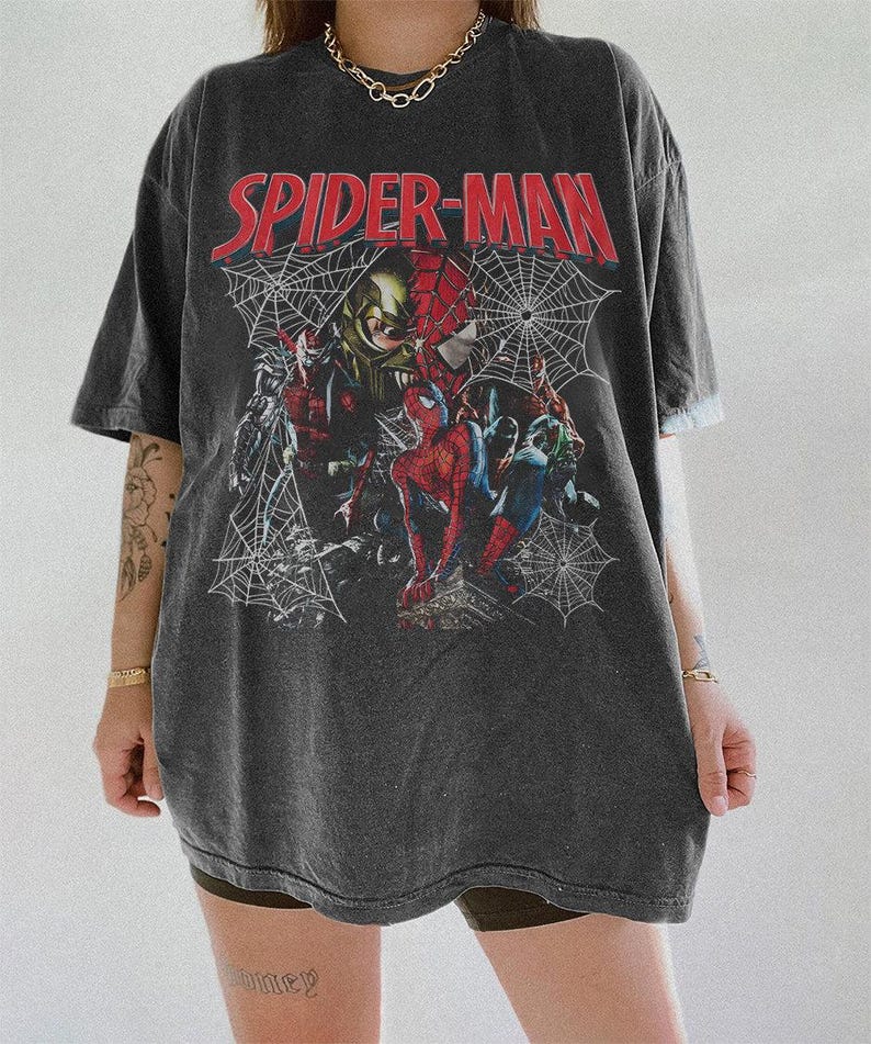 Pode incluir: Uma t-shirt cinzento-carv&atilde;o de corte largo com uma impress&atilde;o gr&aacute;fica do Spider-Man e do Green Goblin. O texto SPIDER-MAN est&aacute; impresso a vermelho no topo, com ilustra&ccedil;&otilde;es de teias de aranha brancas a rodear as personagens na frente.