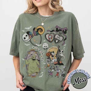 May include: Moss green Comfort Colors t-shirt featuring a graphic design with characters and elements from "The Nightmare Before Christmas." The design includes Jack Skellington, Sally, Oogie Boogie, and other iconic imagery. The shirt has a relaxed fit.