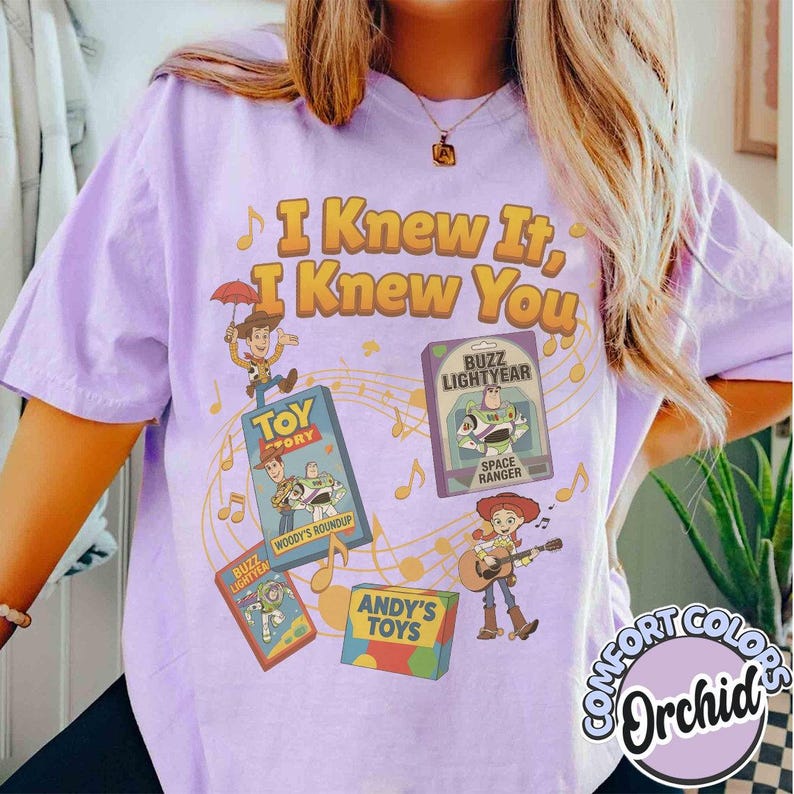 K&ouml;nnte beinhalten: Ein hellviolettes T-Shirt mit dem gelben Schriftzug I Knew It, I Knew You. Die Grafik zeigt Illustrationen von Woody, Buzz Lightyear und Jessie aus Toy Story sowie Noten und Spielzeugschachteln auf einem hellvioletten Hintergrund.