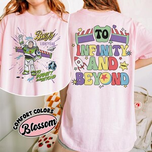 Op de afbeelding: Lichtroze T-shirt met Buzz Lightyear uit Toy Story. De voorkant toont Buzz in zijn ruimtepak met de tekst "Buzz Lightyear" en "To Infinity and Beyond". De achterkant heeft de zin "To Infinity and Beyond" in kleurrijke letters.