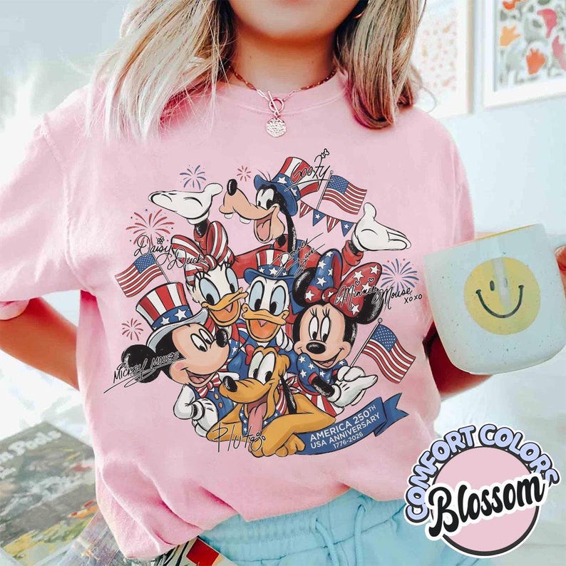 May include: Pink Comfort Colors t-shirt with a graphic of Disney characters Mickey, Minnie, Donald, Daisy, Goofy, and Pluto in patriotic outfits, holding American flags. The design celebrates the America 250th USA Anniversary 1776-2026.