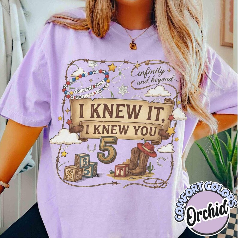 May include: A light purple t-shirt featuring a graphic print with a brown scroll banner that reads I KNEW IT, I KNEW YOU. The design includes a large number 5, a pair of brown cowboy boots, alphabet blocks, a beaded necklace, and clouds.