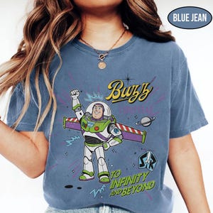 May include: Blue jean-colored t-shirt featuring a graphic of Buzz Lightyear from Toy Story. The design includes the text "Buzz Lightyear" and "To Infinity and Beyond." The shirt has a crew neck and short sleeves.