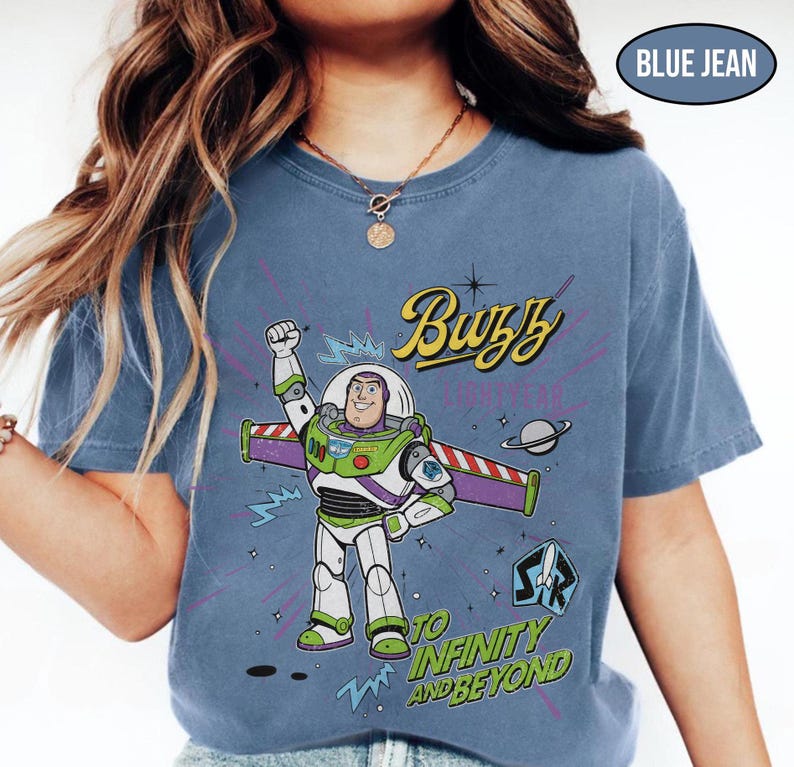 May include: Blue jean-colored t-shirt featuring a graphic of Buzz Lightyear from Toy Story. The design includes the text "Buzz Lightyear" and "To Infinity and Beyond." The shirt has a crew neck and short sleeves.
