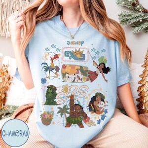 以下が含まれることがあります： ディズニーキャラクターが描かれた水色のTシャツ。モアナ、マウイ、ヘイヘイなどのキャラクターのカラフルなイラストが描かれています。「Disney」の文字が上部にプリントされています。丸首で半袖のTシャツです。
