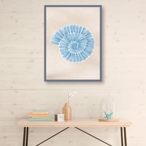 May include: A framed watercolor print featuring a light blue nautilus shell on a beige background. The artwork is encased in a dark blue frame. The image is displayed above a wooden table with decorative items.