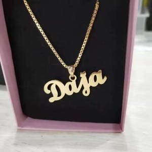 May include: A gold-colored necklace with the name "Daja" in a cursive font. The pendant hangs from a delicate chain. The necklace is displayed inside a pink box with a black interior.