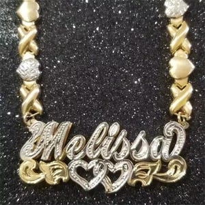 May include: A gold and silver-toned necklace with the name "Melissa" in a cursive font. The necklace features a series of gold X and O links, with heart-shaped and diamond-shaped accents. The bottom of the necklace has heart-shaped designs.
