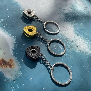 May include: Three metal keychains featuring a rotary engine design. One is silver, one is gold, and one is black. Each keychain has a chain and a silver key ring. The keychains are displayed on a weathered, blue surface.