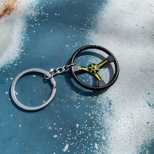 May include: Black and yellow steering wheel keychain. The keychain features a miniature steering wheel design with a black rim and yellow accents. The keychain is attached to a silver key ring and chain.