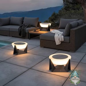 Puede incluir: Lámparas solares para exteriores con un diseño moderno. Las lámparas tienen una parte superior blanca y brillante y una base geométrica negra. La escena incluye muebles de exterior grises y un fondo de montaña, creando un ambiente relajante.